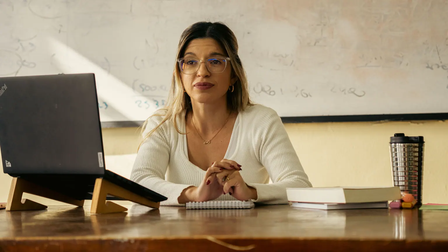 Professora sentada à mesa consulta anotações ao lado de um computador