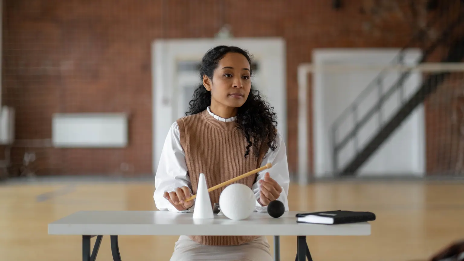 Professora sentada em uma mesa com materiais sonoros em um espaço escolar
