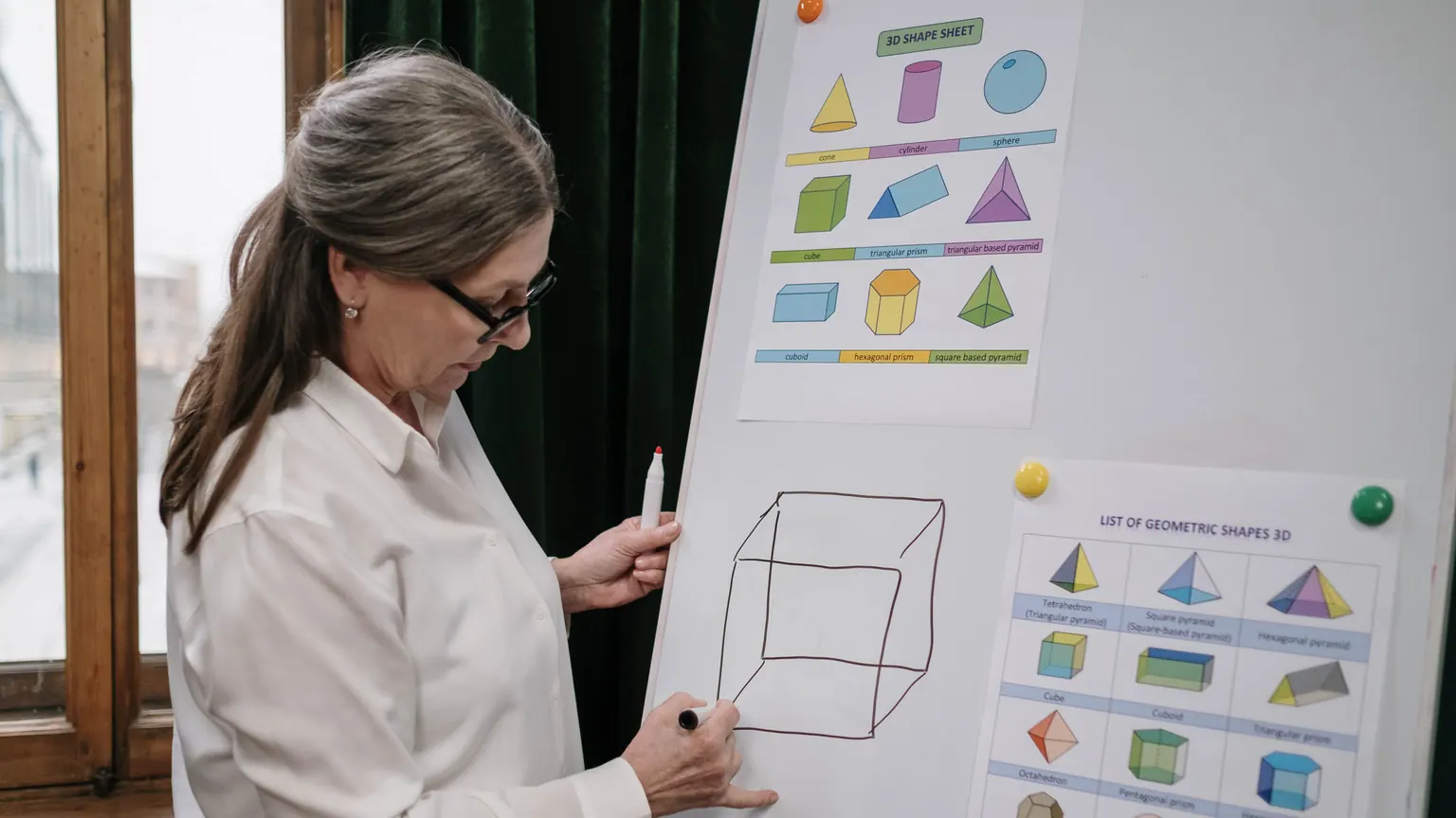 Professora aponta formas geométricas em um quadro de apoio durante aula de matemática