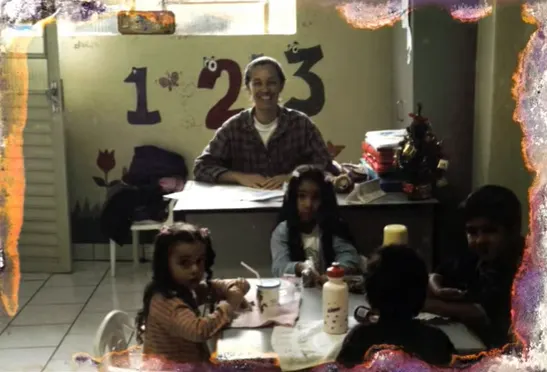 Fotografia antiga: a mãe de Leandro à mesa da escolinha, cercada de crianças fazendo atividades.