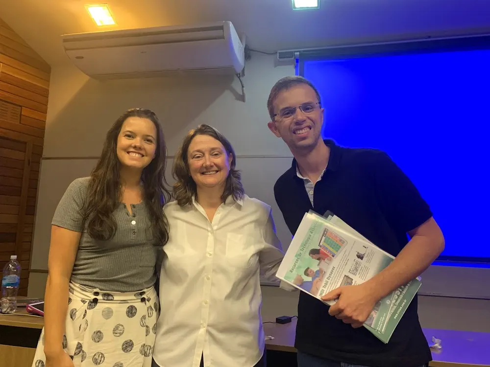 Leandro segurando materiais de comunicação alternativa ao lado da fonoaudióloga Linnea McAfoose, em sala de treinamento.